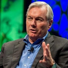 A man in a gray blazer and blue collared shirt sits and speaks.