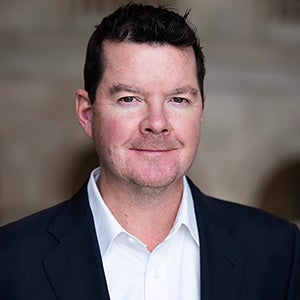 A man wearing a black blazer and a white collared shirt stands and smiles.