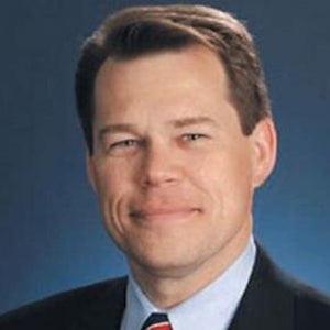 A man wearing a black blazer, light blue collared shirt, and a tie, stands and smiles.