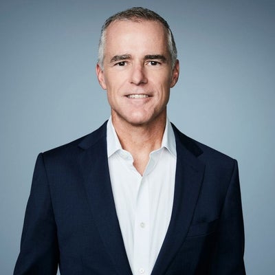 A man in a blue blazer and white collared shirt stands and smiles.