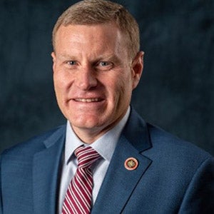 A man in a blue blazer, white collared shirt, and blue and gold striped tie stands inside and smiles.