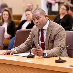 A man in a tan jacket and red necktie sits at a table with a microphone in front of him.