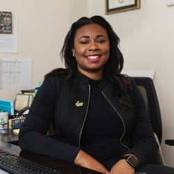 A woman in a black outfit sits at a desk facing the camera.