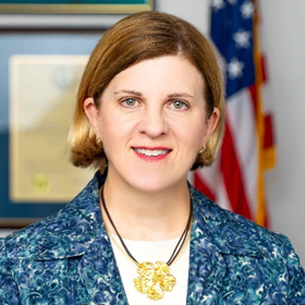 A woman in a blue and white blazer and white shirt stands smiling in front of an American flag