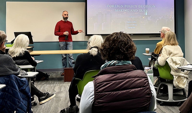 A man with a long beard and wearing a red sweater stands in front of a class of students.