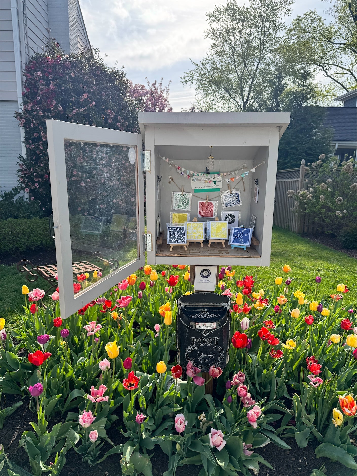 9 art pieces in the Free Little Art Gallery. The gallery is surrounded by tulips and has a black container under it that says "Free Art Kits."