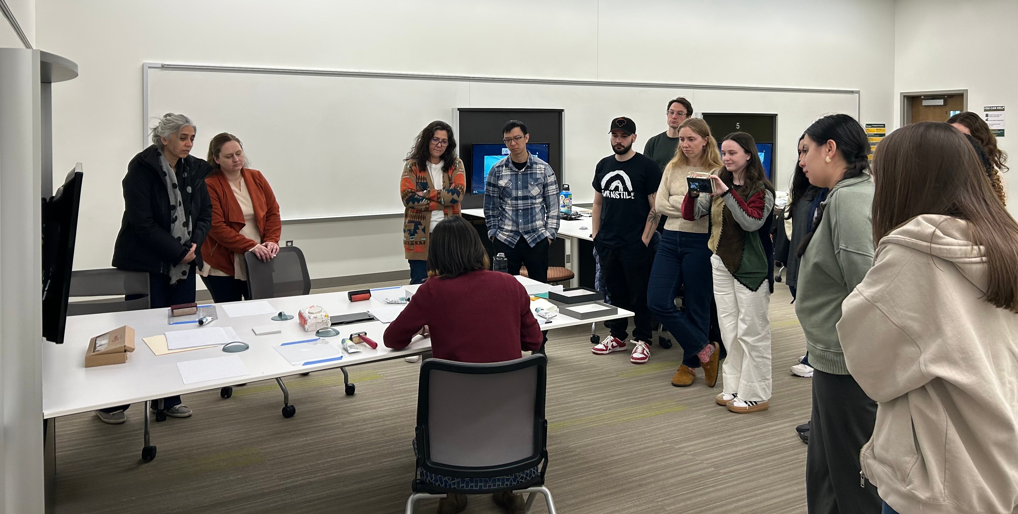 13 students are standing arounda table where the professor is demonstrating printmaking techniques. The professor is sitting and has her back turned from the camera.