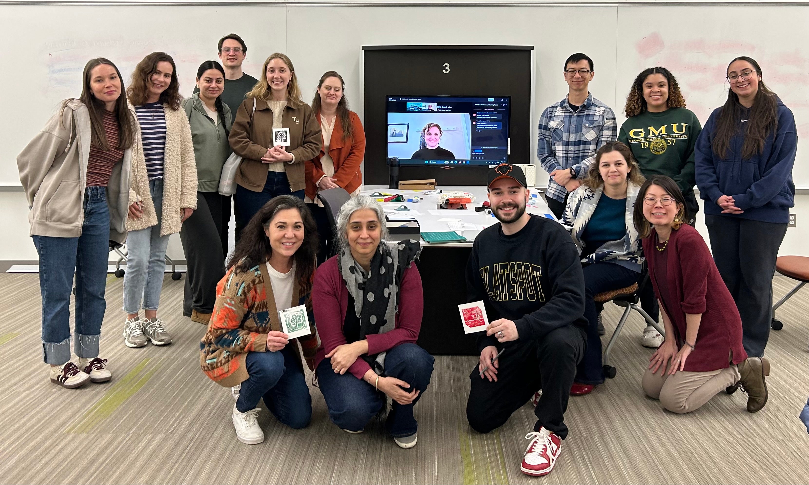 13 students in SOCW 689 standing next to each other or kneeling and smiling to the camera. Professor Goerdt is kneeling and smiling at the camera while Stacey Schwartz is on the tv screen smiling at the camera. 3 students are holding up mini art prints.