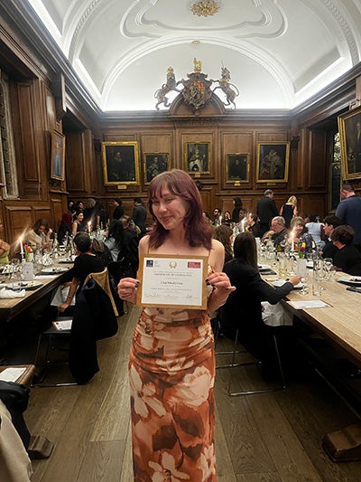 Lisa Misaki Gray dressed in formal attire and holding a certificate of completion in a dining hall