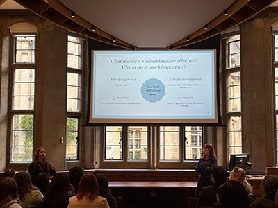 Gray and Gizzi speaking to a room of attendees in front of a presentation slide entitled "what makes a silence breaker effective. Why is the work important?"