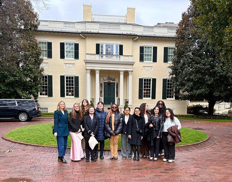 A group of women stand in front of a yellow brick building with white columns.