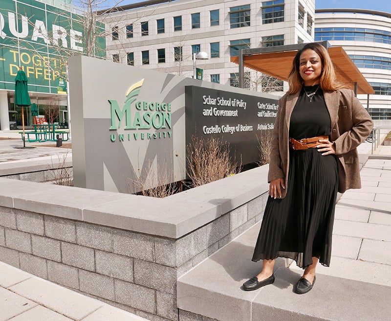 A woman with light brown hair in a black dress stands in front of a granite sign for a university.