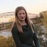 Portrait of Abbi Tesreau, a young woman with long blonde hair wearing a dark green top, standing outdoors near a river with a bridge in the background.