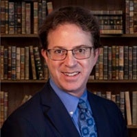 Professional headshot of Mark S. Zaid wearing a blue suit, blue shirt, and patterned tie, standing in front of bookshelves.