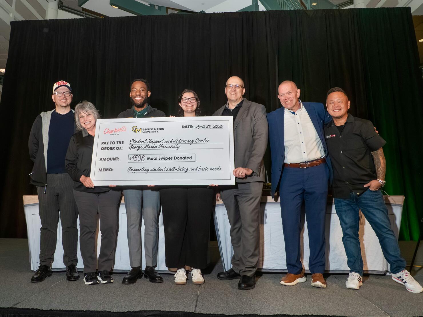 Chef Jet Tila helps George Mason staff hold a large check on stage showing meal swipes donated