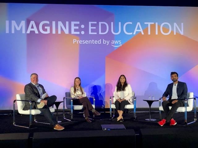 Maya Chatterjee sitting in a row with other AWS Imagine EDU student speakers in front of a colorful screen backdrop.