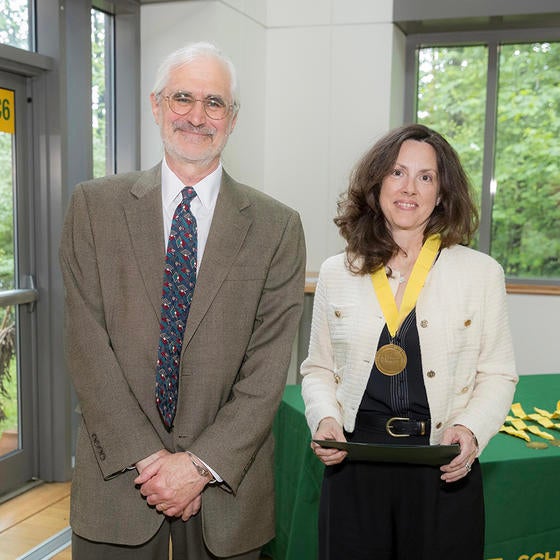 A man in a tan jacket stands next to a woman in a white jacket wearing a medal.