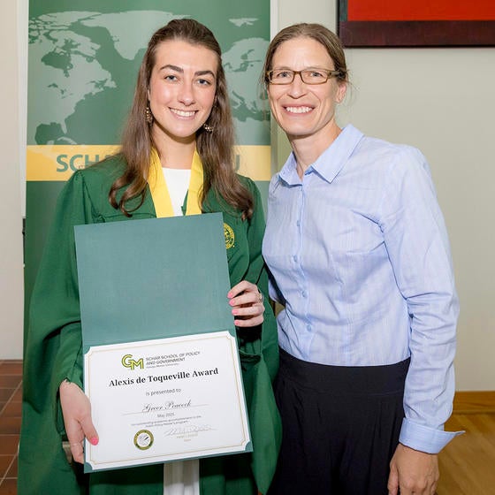 A woman in a green gown holds a certificate while a woman in a blue shirt smiles.
