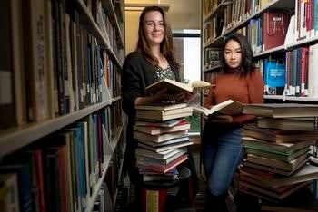 Two women standing in the Mason library stacks with a cart of books and books open in their hands, looking at the camera. 