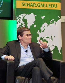 A man in a suit with a microphone on his white shirt sits in a leather chair in front of a banner.