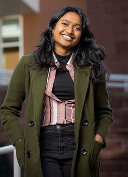Schar School student Ruthu Josyula standing and smiling in front of a brick building while wearing a green jacket
