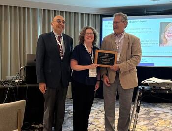 Three people in a conference room, where the person in the middle is receiving a plaque