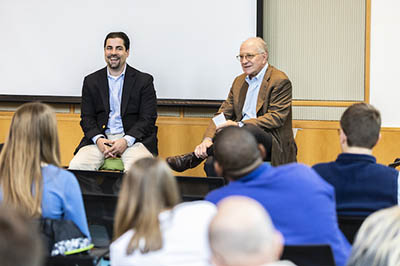 Two men, one laughing, sit in seats in front of a small audience.