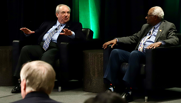 Two men in suits sit in chairs in front of a green curtain.