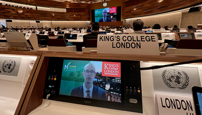 Two video screens, one large in the back and one small in the front, are seen in a large conference room with a stylized ceiling.