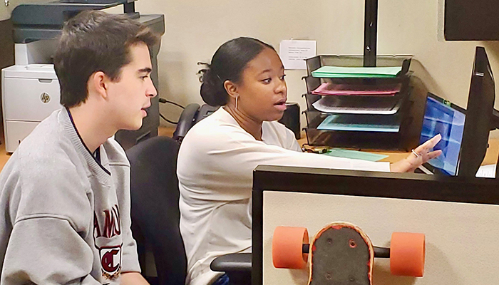 A woman gestures at a computer while a man sits in a chair next to her desk.