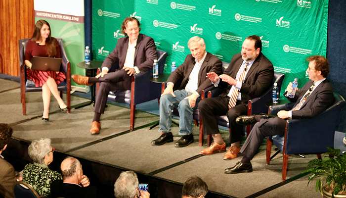 Five people, including a woman in a maroon dress, and four men in dark suits sit in chairs on a stage.