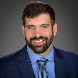 A man with dark hair and beard in a suit jacket and tie smiles at the camera.