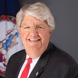 A man in a dark jacket and red necktie stands in front of a Virginia state flag.