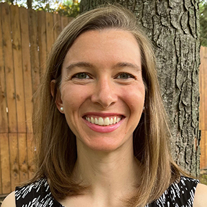 A woman in a black and white top stands in front of a tree and smiles.