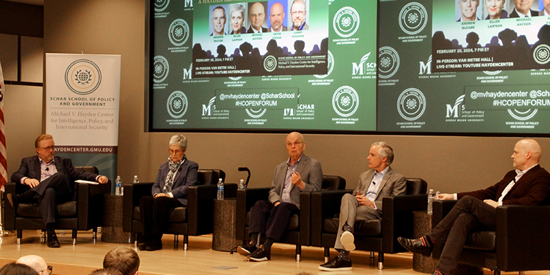 Four men and a woman sit in black chairs on a stage with a green backdrop.