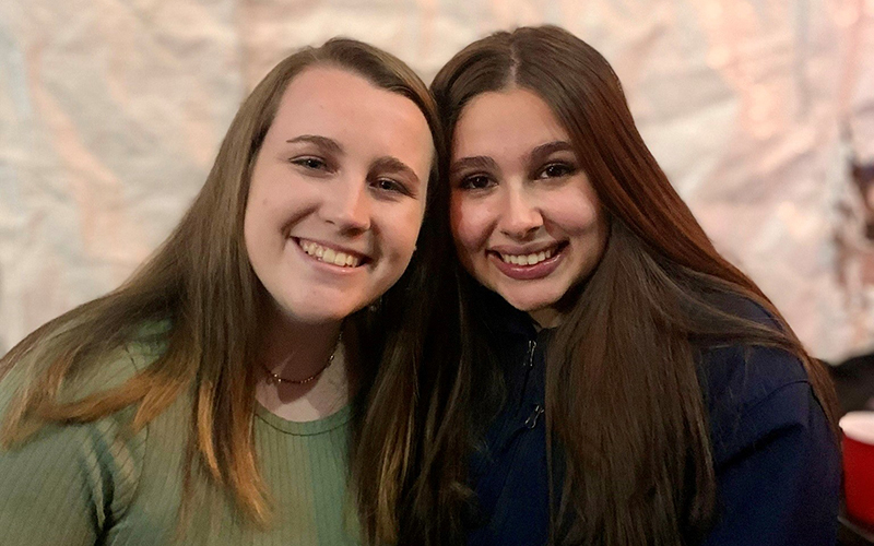 Two young adult women with long hair sit close to each other and smile at the camera.