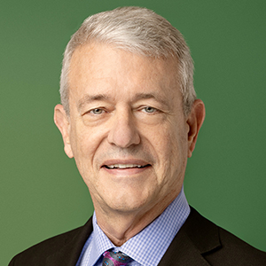 Phillip Mink sits in front of a green background wearing a black jacket, blue collared shirt, and a tie while smiling.