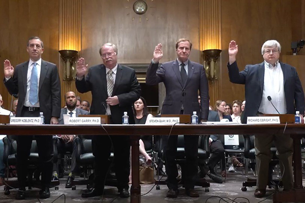 Four men raise their right hands as they stand in front of a table with nameplates and microphones.