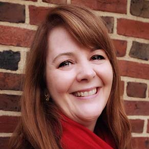 A woman with red hair and wearing a red scarf smiles at the camera in front of a brick wall.