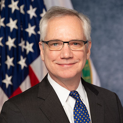 A man in a dark suit, white shirt, and blue tie stands smiling in front of an American flag