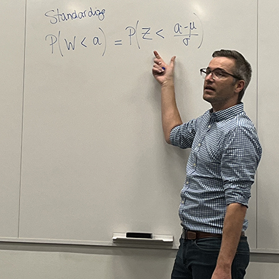 A man wearing glasses and a blue checked button-down shirt with the sleeves rolled up gestures toward a white board at the front of a classroom.
