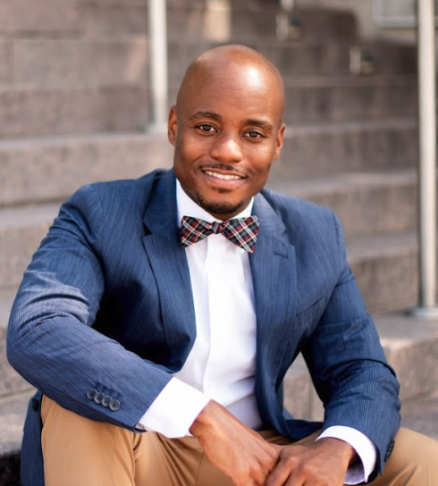 A man sitting in front of concrete stairs while wearing a blue suit with a white shirt. He is smiling at the camera while his arms are leaning against his legs and holding his hands together.