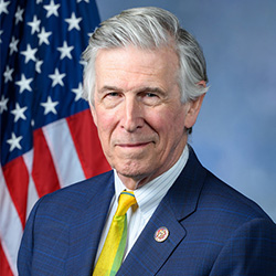 A man in a blue blazer, white collared shirt, and yellow tie stands in front of an American flag and smiles.