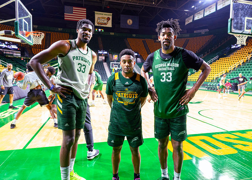 Three men in basketball jerseys pose for the camera while others play basketball behind them.