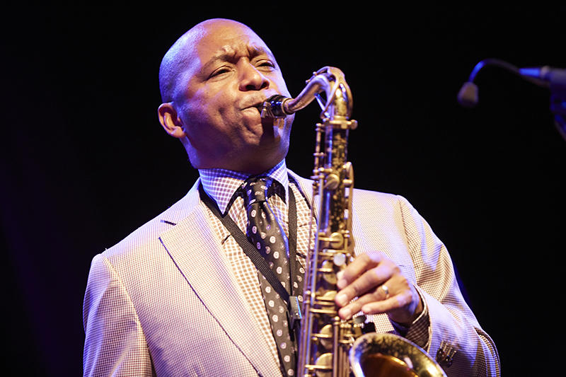 Branford Marsalis leans his head back, playing an alto saxophone and wearing a beige suit and polka-dot tie.