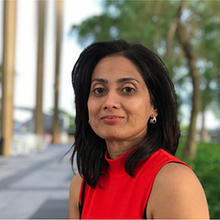 A woman with long black hair and a red shirt stands outside and smiles.