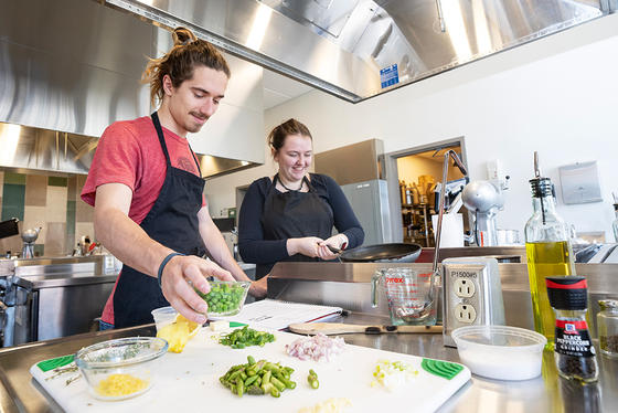Two students stand behind a stove. One is stirring the recipe cooking in a pan, while the other prepares to add an ingredient to the pan.