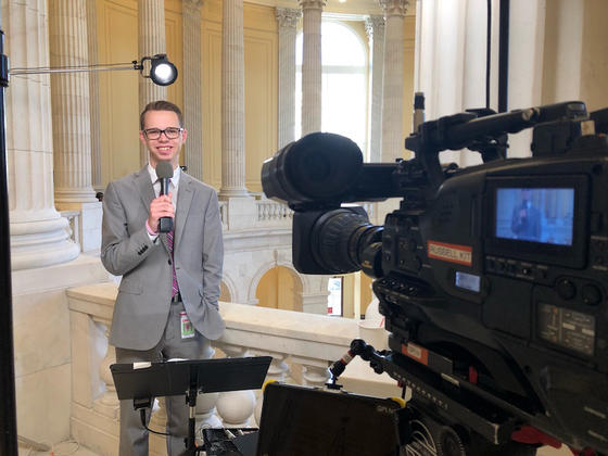 Nathan Crawford stands at the balcony of the capitol rotunda holding a microphone and looking into the camera. He is smiling and wearing a gray business suit.