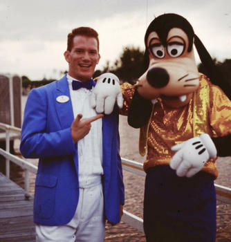 Violinist Peter Wilson in a blue sportcoat, white shirt, and black bowtie, who now serves as the conductor of the American Festival Pops Orchestra, poses with the character Goofy at Disney World.