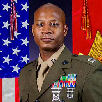 A man in a green military jacket gazes at the camera with colorful flags behind him.